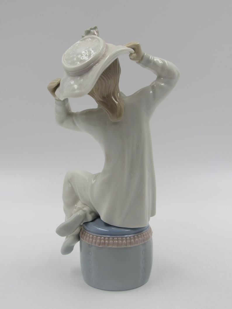 May include: A porcelain figurine of a girl wearing a white dress and a white hat with a pink ribbon. She is sitting on a blue and pink stool.