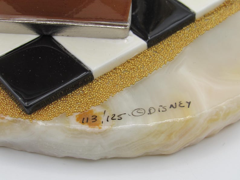 K&ouml;nnte beinhalten: Ein dekoratives Kunstwerk mit geometrischem Design. Das St&uuml;ck zeigt schwarze, wei&szlig;e und braune Fliesen, akzentuiert mit goldfarbenen Perlen. Die Basis ist ein marmorierter, cremefarbener Stein. Der Text "113/125 &copy; Disney" ist sichtbar.