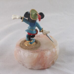 K&ouml;nnte beinhalten: Eine Figur von Mickey Mouse im Robin Hood-Kost&uuml;m, die einen Dolch h&auml;lt. Die Figur steht auf einem rosa-wei&szlig;en Steinsockel mit goldenen Akzenten. Mickey tr&auml;gt eine blaue Tunika, rote Shorts und einen gr&uuml;nen Hut mit roter Feder.