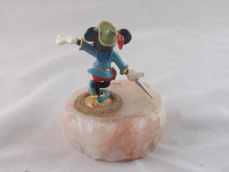 K&ouml;nnte beinhalten: Eine Figur von Mickey Mouse im Robin Hood-Kost&uuml;m, die einen Dolch h&auml;lt. Die Figur steht auf einem rosa-wei&szlig;en Steinsockel mit goldenen Akzenten. Mickey tr&auml;gt eine blaue Tunika, rote Shorts und einen gr&uuml;nen Hut mit roter Feder.