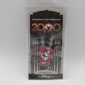 Könnte beinhalten: Ein Disney Store Pin zum Millennium, mit Minnie Mouse. Der Pin ist rot, schwarz und weiß, mit dem Text "Countdown to the Millennium 2000" und "99 Pins to Go". Der Pin ist in einer transparenten Plastiktüte verpackt.