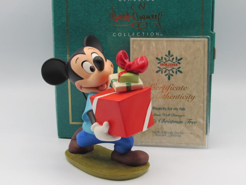 May include: A Mickey Mouse figurine from the Disney Classics Collection, holding a large red gift box and smaller presents. The figurine is wearing blue pants and a light blue shirt. A certificate of authenticity is visible in the background.