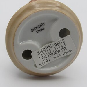 Op de afbeelding: Een keramieken beeldje van een brandweerman, met een beige en wit kleurenschema. De basis van het beeldje heeft twee gaten en een sticker met de tekst "MM FIREMAN FIG" en "MADE IN CHINA". Het Disney-logo is ook zichtbaar.