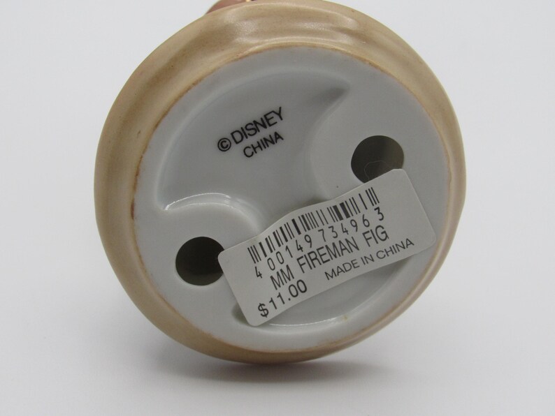 Op de afbeelding: Een keramieken beeldje van een brandweerman, met een beige en wit kleurenschema. De basis van het beeldje heeft twee gaten en een sticker met de tekst "MM FIREMAN FIG" en "MADE IN CHINA". Het Disney-logo is ook zichtbaar.