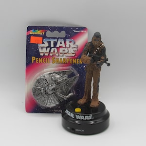 Puede incluir: Un sacapuntas de Star Wars con una figura de Chewbacca sobre una base negra. El embalaje incluye un sacapuntas del Halcón Milenario y las palabras "Star Wars Pencil Sharpener".