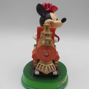 May include: A Mickey Mouse figurine in a conductor's outfit, riding a red and gold train. The figurine is on a green base with the text "NEW MICKEY". The train has a round front and gold accents.