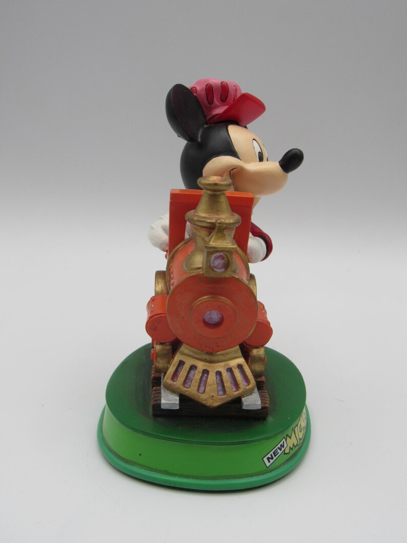 May include: A Mickey Mouse figurine in a conductor's outfit, riding a red and gold train. The figurine is on a green base with the text "NEW MICKEY". The train has a round front and gold accents.