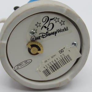 May include: A white ceramic figurine base with a gold-colored winding mechanism. The base has a sticker that reads "25 Walt Disney World" and "Crafted in Sri Lanka". A price tag with a barcode is also attached.