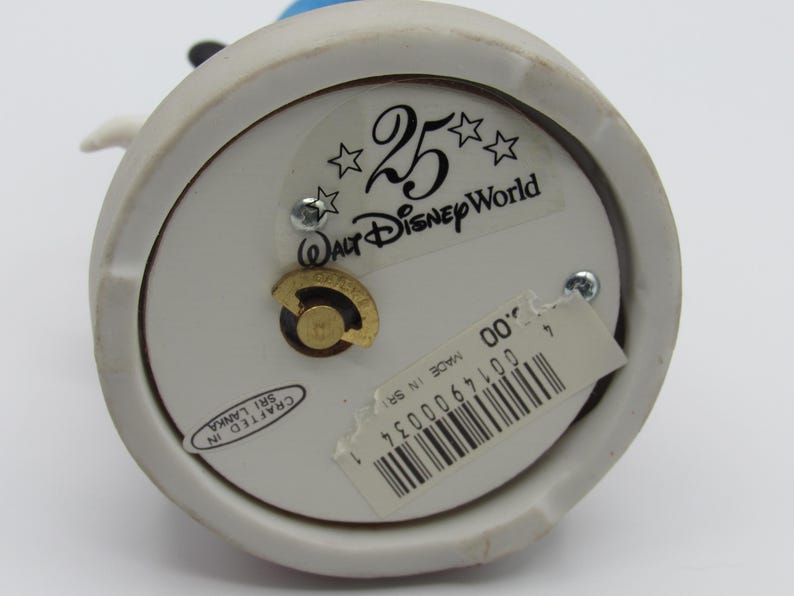 May include: A white ceramic figurine base with a gold-colored winding mechanism. The base has a sticker that reads "25 Walt Disney World" and "Crafted in Sri Lanka". A price tag with a barcode is also attached.