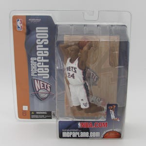 May include: A McFarlane Sports Picks NBA action figure of Richard Jefferson in a white New Jersey Nets jersey with red and blue accents. The figure is posed in a mid-air dunk, holding a basketball. The figure is packaged in a clear plastic blister pack with a cardboard backing. The backing features a photo of Jefferson, the Nets logo, and the text "Richard Jefferson Nets NBA.com mcfarlane.com".