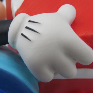 May include: Close-up of a white cartoon glove with three black lines on the palm. The glove is part of a larger figure, with a black sleeve and blue and red elements visible.
