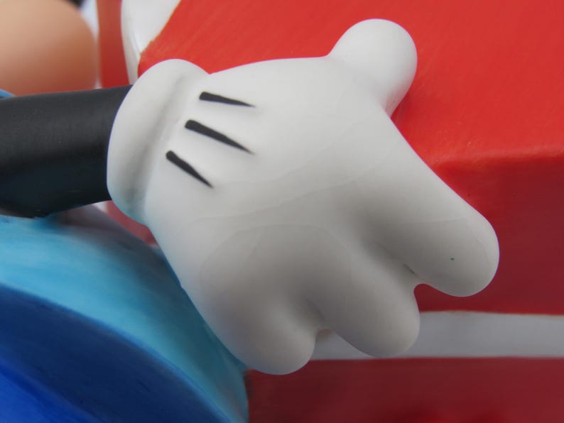 May include: Close-up of a white cartoon glove with three black lines on the palm. The glove is part of a larger figure, with a black sleeve and blue and red elements visible.