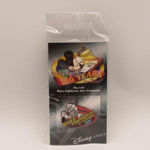 Puede incluir: Un juego de pines de Disney en un paquete de plástico transparente. El pin superior presenta a Mickey Mouse y Goofy con el texto "100 Years of Dreams". El pin inferior tiene a Buzz Lightyear con el texto "Buzz Lightyear 2000". El paquete dice "Disney store".