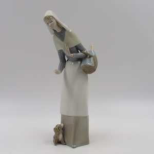 May include: A ceramic figurine depicting a woman in a long white dress and headscarf, holding a basket. A small dog is at her feet, gazing upwards. The figurine is painted in muted tones of white, grey, and brown.
