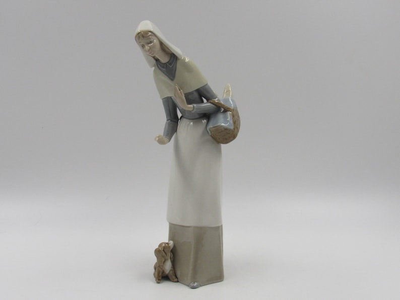 May include: A ceramic figurine depicting a woman in a long white dress and headscarf, holding a basket. A small dog is at her feet, gazing upwards. The figurine is painted in muted tones of white, grey, and brown.