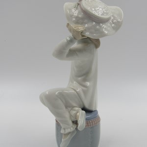 May include: A porcelain figurine of a girl wearing a white dress and a large white hat with pink trim. She is sitting on a blue and white base.