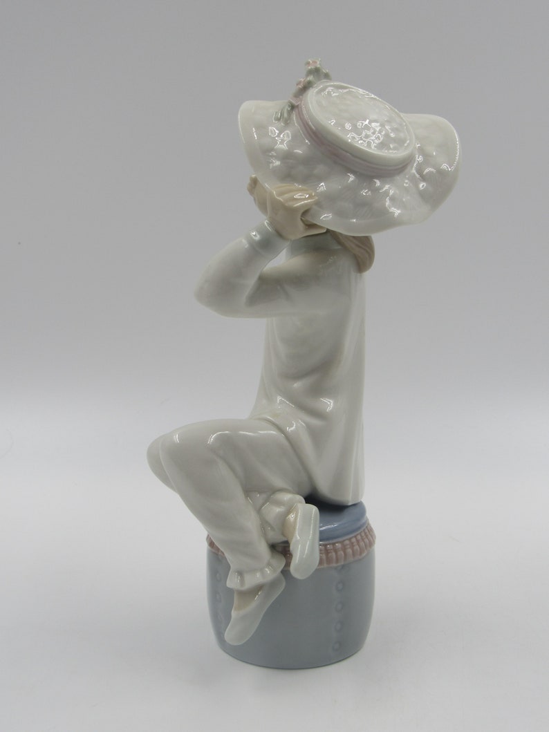 May include: A porcelain figurine of a girl wearing a white dress and a large white hat with pink trim. She is sitting on a blue and white base.