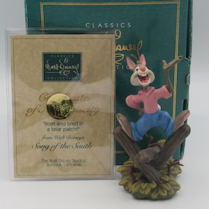 Puede incluir: Una figura de Disney Classics de Br'er Rabbit de "Canción del Sur", con camisa rosa y pantalones azules, posada en una valla de madera. Incluye un certificado de autenticidad y una caja verde.