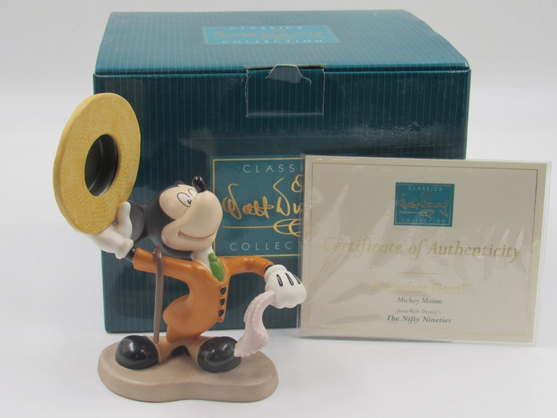 May include: A Mickey Mouse figurine from the Disney Classics Collection, holding a hat and cane, dressed in an orange suit with a green tie. The figurine is displayed with its original box and certificate of authenticity.