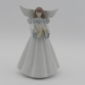May include: A porcelain angel figurine with wings, a white dress, and brown hair. The angel is holding a small, round, white object in her hands.