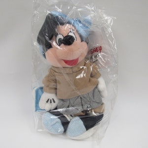May include: A plush Mickey Mouse toy wearing a blue hat, brown jacket, gray pants, and blue shoes. The toy is packaged in a clear plastic bag with a Disney label.