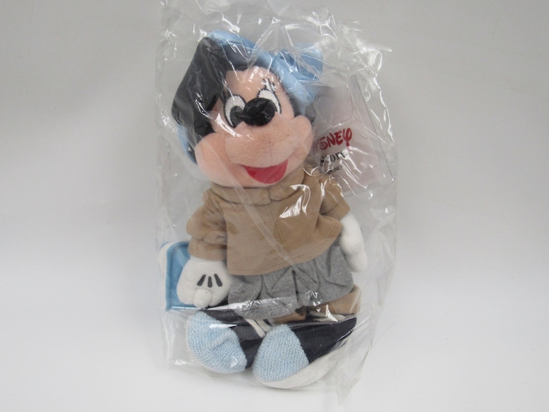 May include: A plush Mickey Mouse toy wearing a blue hat, brown jacket, gray pants, and blue shoes. The toy is packaged in a clear plastic bag with a Disney label.