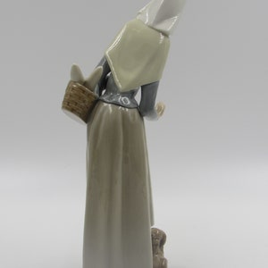 May include: A ceramic figurine of a woman in a long dress and head covering, carrying a basket. The figure is in shades of grey, beige, and white, with a small dog at her feet. The figurine has a glossy finish.