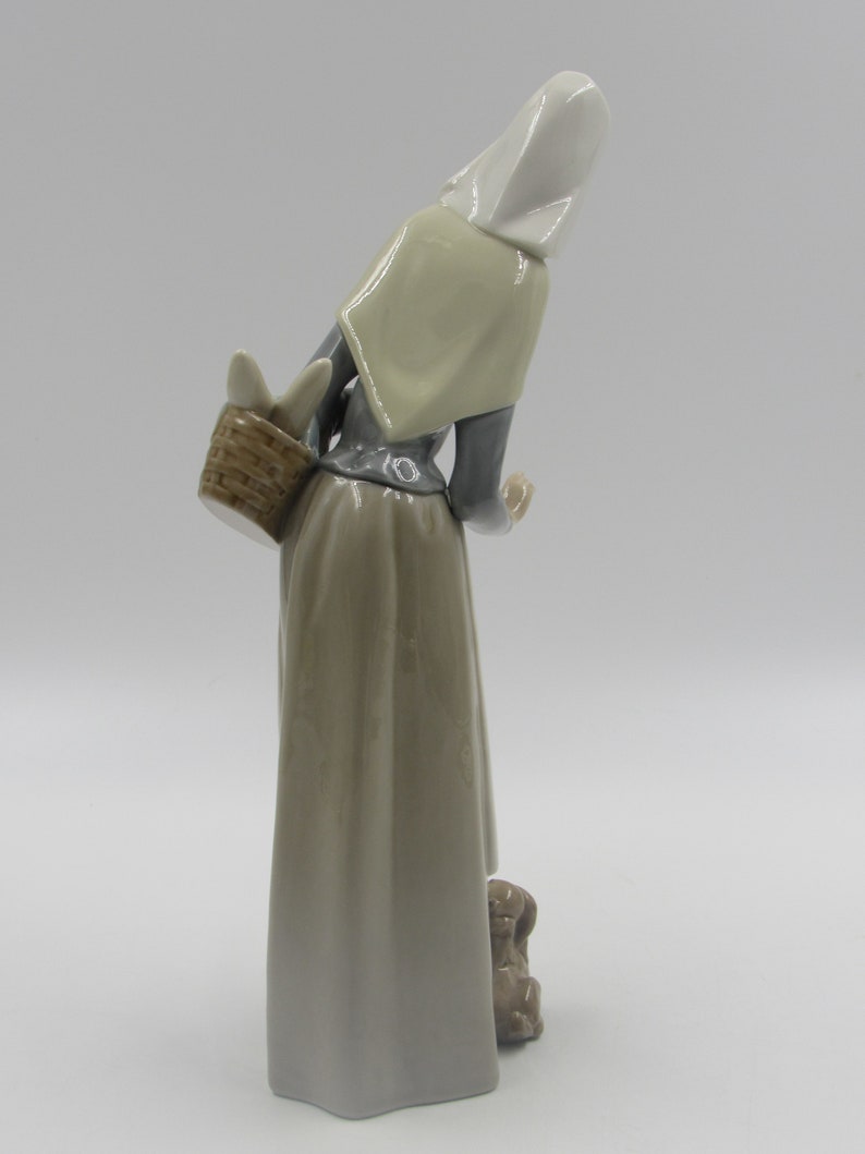 May include: A ceramic figurine of a woman in a long dress and head covering, carrying a basket. The figure is in shades of grey, beige, and white, with a small dog at her feet. The figurine has a glossy finish.