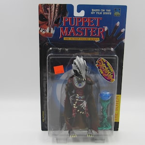 May include: A plastic package containing a Puppet Master action figure of The Totem. The figure is wearing a red and black robe with a silver helmet. The figure is holding a blue and green scepter. The package is labeled "Puppet Master" and "The Totem".