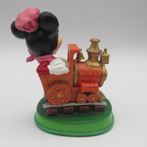 May include: A decorative figurine of Minnie Mouse driving an orange and gold train. Minnie is wearing a pink hat, scarf, and red jacket. The train sits on a green base with brown tracks. The figurine is a collectible item.