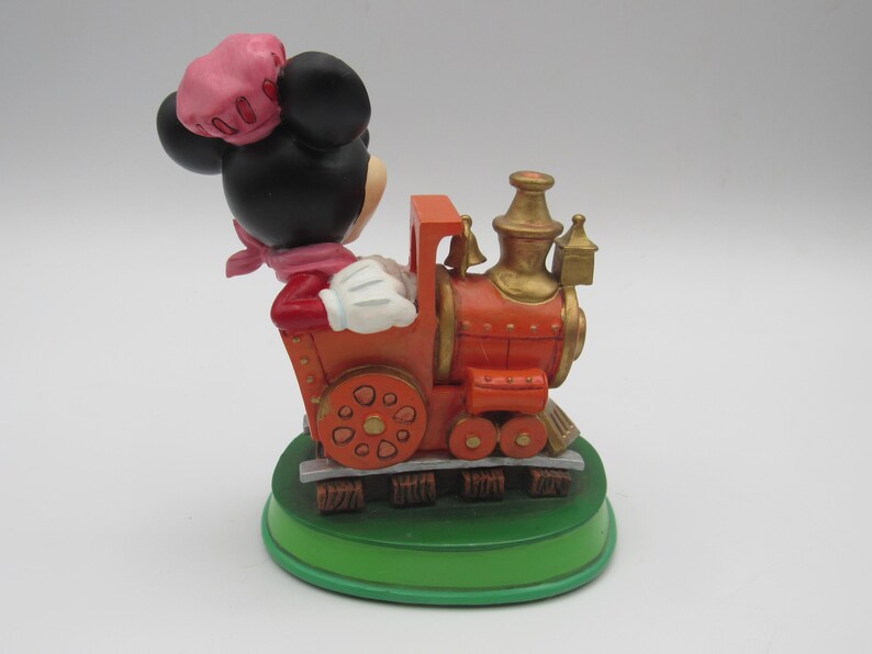 May include: A decorative figurine of Minnie Mouse driving an orange and gold train. Minnie is wearing a pink hat, scarf, and red jacket. The train sits on a green base with brown tracks. The figurine is a collectible item.