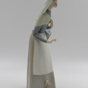 May include: A ceramic figurine depicting a woman in a long dress and head covering, accompanied by a small dog. The woman is in a walking pose, with a neutral expression. The figurine is predominantly white, grey, and beige.