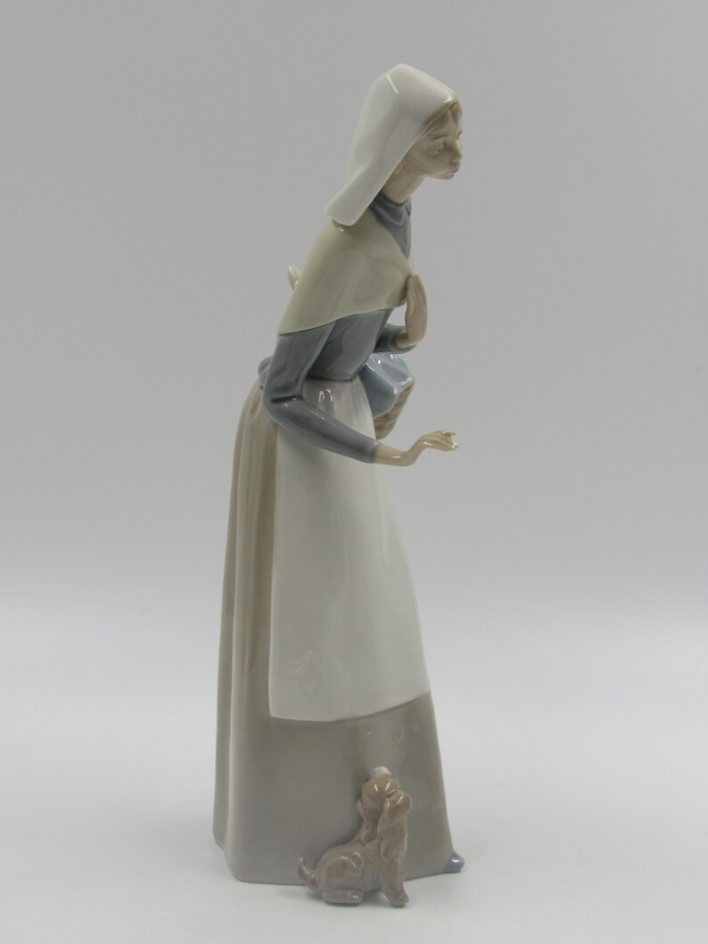 May include: A ceramic figurine depicting a woman in a long dress and head covering, accompanied by a small dog. The woman is in a walking pose, with a neutral expression. The figurine is predominantly white, grey, and beige.