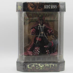 Könnte beinhalten: Eine Burnt Spawn Actionfigur in einer sechseckigen Kunststoff-Verpackung. Die Figur trägt einen schwarzen Anzug mit roten Akzenten, silbernen Ketten und stacheligen Handschuhen. Ein kleines graues wolfsähnliches Wesen steht neben der Figur. Die Verpackung zeigt den Text Burnt Spawn und Spawn.