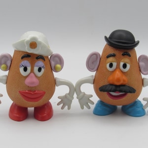 Può includere: Due giocattoli di Mr. Potato Head. Uno ha un cappello bianco e scarpe rosse, l'altro un cappello nero e scarpe blu. Entrambi hanno braccia, orecchie e lineamenti del viso.