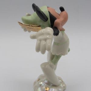 Op de afbeelding: Een porseleinen beeldje van een stripfiguur, met een groene hoed, zwarte oren en een gouden veer. Het personage draagt een witte outfit en staat op een wolkenachtige basis met een gouden ster. Het beeldje heeft een parelmoer afwerking.