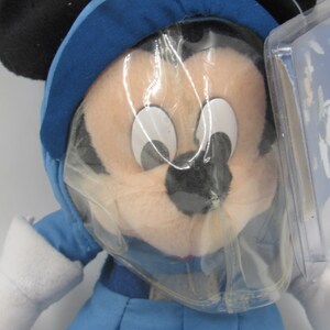 May include: A plush Mickey Mouse doll wearing a blue winter outfit with a hood and white gloves. The doll has black ears and is partially covered in plastic packaging. A tag is attached.
