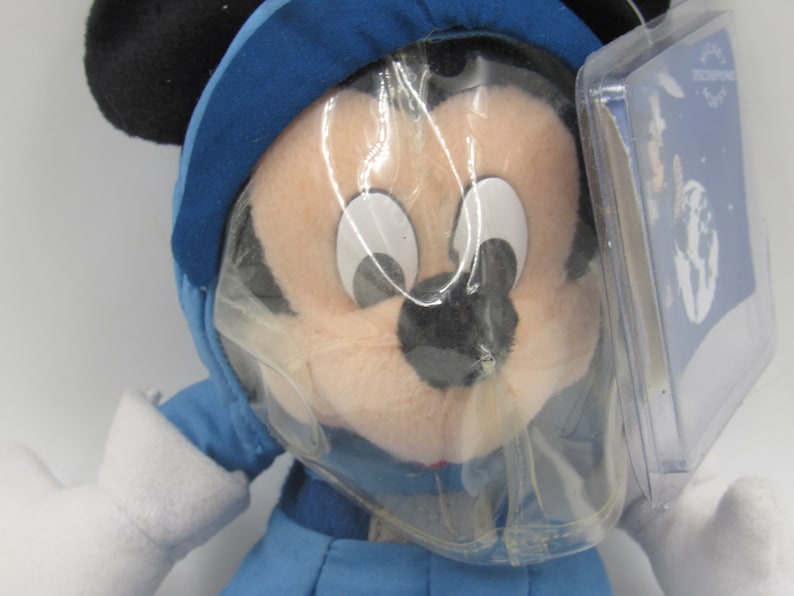 May include: A plush Mickey Mouse doll wearing a blue winter outfit with a hood and white gloves. The doll has black ears and is partially covered in plastic packaging. A tag is attached.