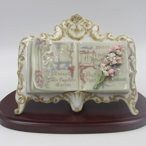 May include: A porcelain figurine of an open book with a bouquet of pink flowers on the right page. The book is on a stand with a wood base. The book's pages have text that reads "Scene II The Capulet's Garden" and "Romeo, if just that what thou art, But light..."