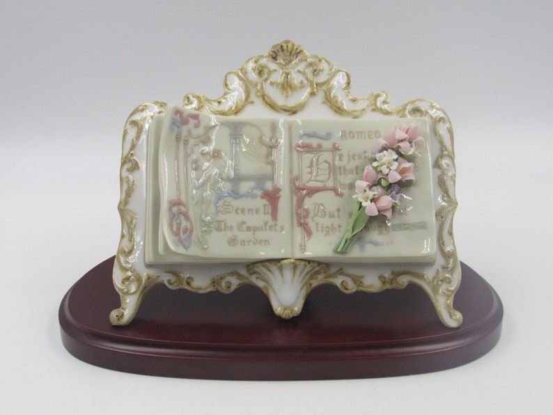 May include: A porcelain figurine of an open book with a bouquet of pink flowers on the right page. The book is on a stand with a wood base. The book's pages have text that reads "Scene II The Capulet's Garden" and "Romeo, if just that what thou art, But light..."