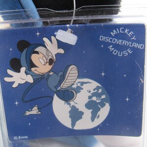 May include: A blue tag with the words "MICKEY DISCOVERYLAND MOUSE" and a cartoon image of Mickey Mouse in a space suit. Mickey is depicted floating in space with a planet and a spaceship in the background. The tag is in a clear plastic case.