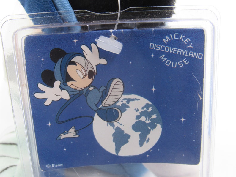 May include: A blue tag with the words "MICKEY DISCOVERYLAND MOUSE" and a cartoon image of Mickey Mouse in a space suit. Mickey is depicted floating in space with a planet and a spaceship in the background. The tag is in a clear plastic case.
