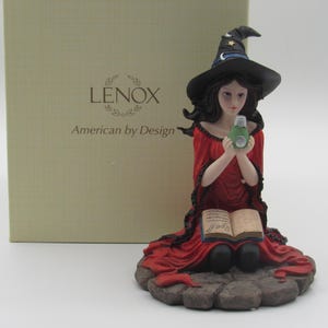 Könnte beinhalten: Eine Lenox-Figur einer Hexe in einem roten Kleid und einem schwarzen Hut, die eine grüne Zaubertrankflasche hält. Die Figur sitzt auf einem Steinsockel mit einem offenen Buch. Die Schachtel hat das Lenox-Logo und den Text "American by Design".