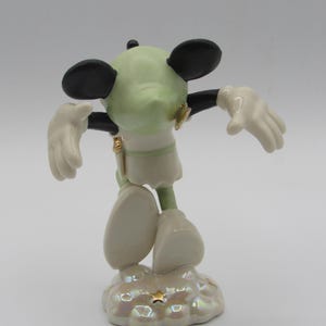 Op de afbeelding: Een keramieken beeldje van een muisachtig personage met een lichtgroen hoofd, zwarte oren en witte handschoenen en schoenen. Het figuur staat op een wolkvormige basis met gouden accenten.