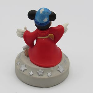 May include: A figurine of a cartoon mouse wearing a blue wizard hat with white stars and a red robe. The mouse is standing on a gray base decorated with silver stars. The mouse is pointing with its white gloved hands.