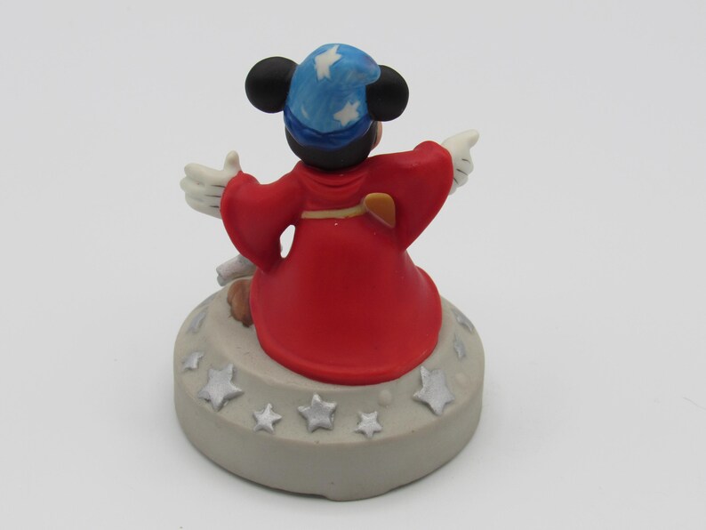 May include: A figurine of a cartoon mouse wearing a blue wizard hat with white stars and a red robe. The mouse is standing on a gray base decorated with silver stars. The mouse is pointing with its white gloved hands.