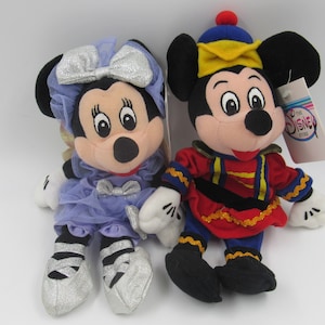 May include: A pair of plush Mickey Mouse and Minnie Mouse toys. Minnie is wearing a purple dress and silver shoes. Mickey is dressed as a nutcracker in a red and blue costume. The toys have The Disney Store tags attached.