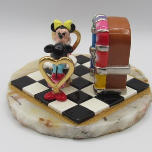 K&ouml;nnte beinhalten: Dekorative Figur mit Minnie Maus und einer Jukebox. Minnie Maus befindet sich in einem goldfarbenen herzf&ouml;rmigen Rahmen und tr&auml;gt eine gelbe Schleife, rote Schuhe und blaue Shorts. Die Jukebox ist braun mit bunten Akzenten, auf einer schwarz-wei&szlig;en karierten Basis.
