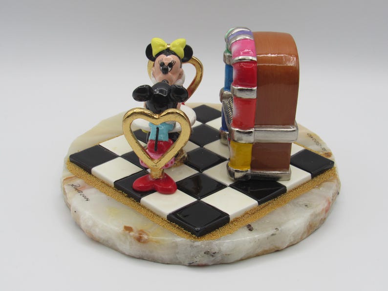 K&ouml;nnte beinhalten: Dekorative Figur mit Minnie Maus und einer Jukebox. Minnie Maus befindet sich in einem goldfarbenen herzf&ouml;rmigen Rahmen und tr&auml;gt eine gelbe Schleife, rote Schuhe und blaue Shorts. Die Jukebox ist braun mit bunten Akzenten, auf einer schwarz-wei&szlig;en karierten Basis.