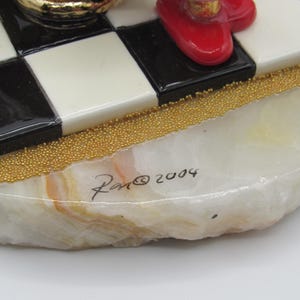 K&ouml;nnte beinhalten: Dekorative Figurenbasis mit einem schwarz-wei&szlig;en Schachbrettmuster. Die Basis besteht aus einem hellfarbigen Stein mit goldfarbenen Perlenakzenten. Die Signatur "Ron &copy; 2004" ist auf der Basis sichtbar.